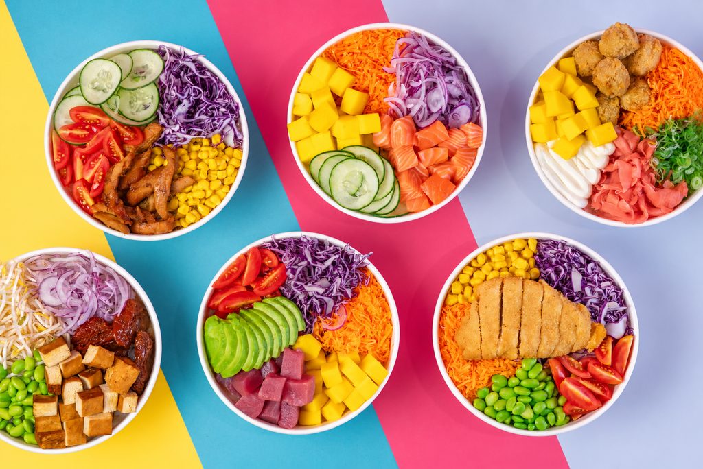 Seis bowls de poke vistos de cima, sobre faixas amarelas, azuis, rosa e lilás: salmão, atum, abacate, manga, edamame, couve roxa e frango panado.