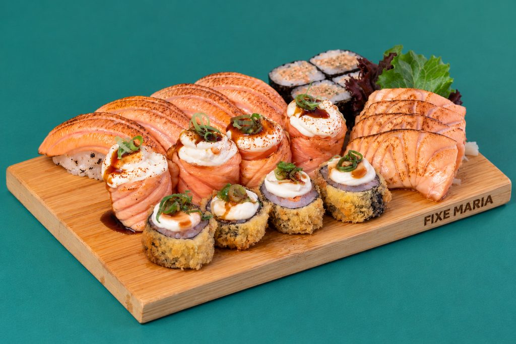 Tábua de madeira com gunkan de salmão braseado, nigiri e hot rolls, sobre fundo verde-azulado.