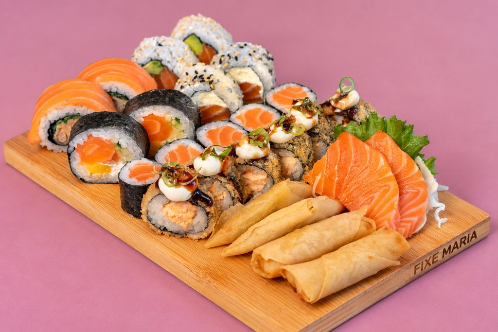 Tábua mista com uramaki, hossomaki, sashimi de salmão e crepes fritos, sobre fundo malva.