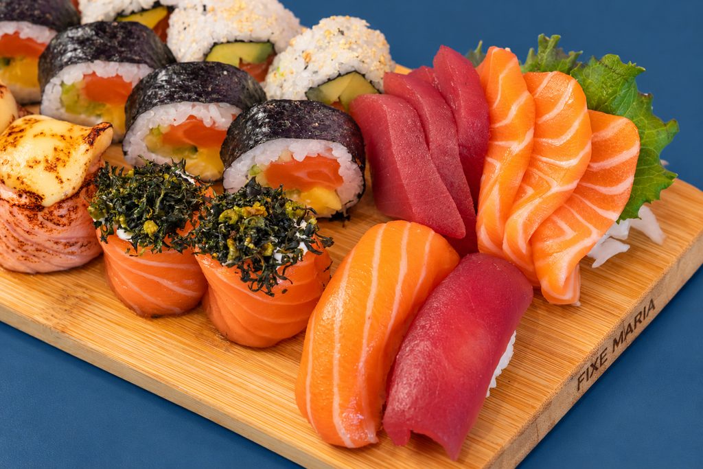 Tábua de madeira com sashimi e nigiri de salmão e atum, uramaki e gunkan, sobre fundo azul-marinho.